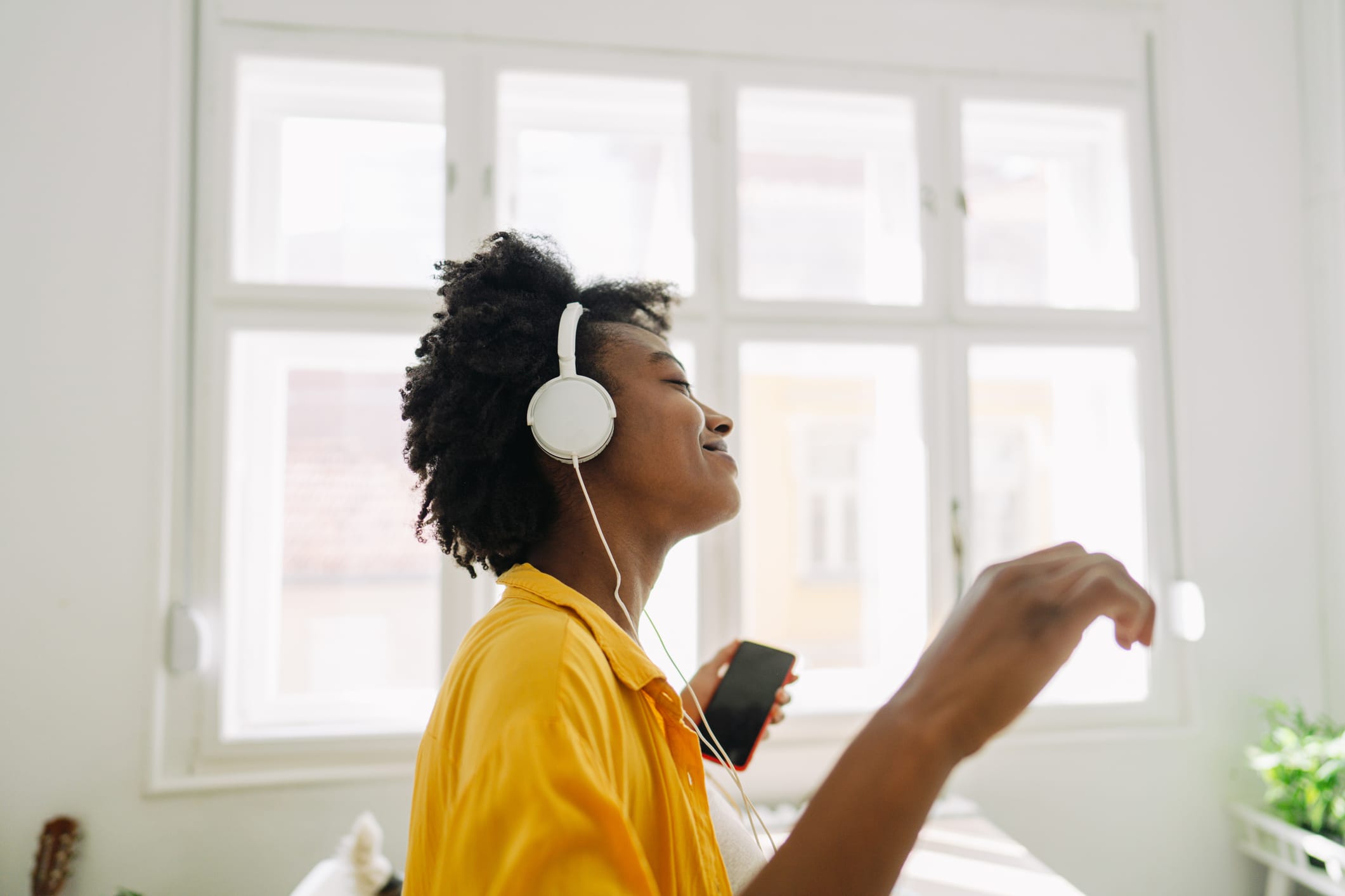 Photo of a young woman of African American ethnicity, using mobile phone and listening to some good music, in a living room